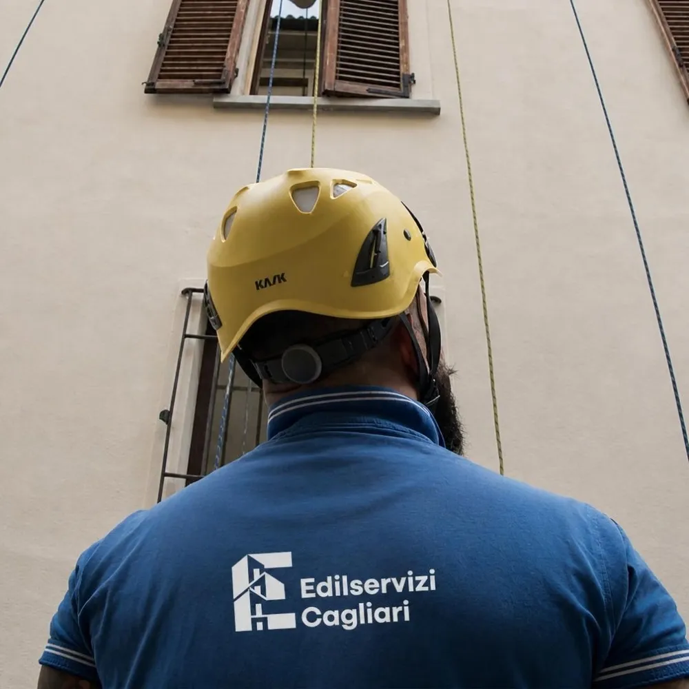 An Edilservizi Cagliari worker in a helmet and company polo shirt, seen from behind in front of a façade under repair